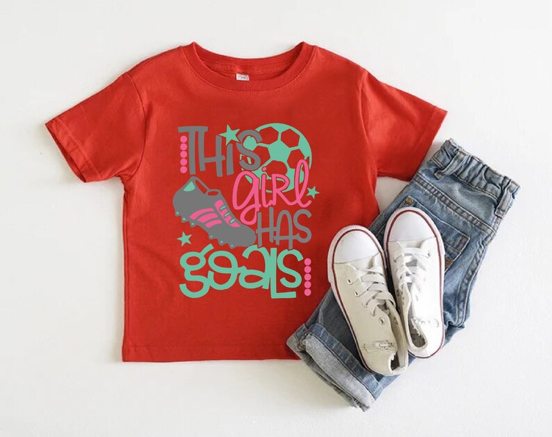 May include: Red short-sleeved t-shirt with the text "This girl has goals" in green and pink lettering. A soccer ball and a soccer cleat are also featured on the shirt.