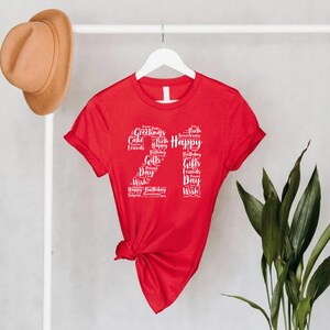 May include: Red short-sleeved t-shirt with a white graphic of the number 27 made up of words related to birthdays, such as "Happy", "Birthday", "Cake", "Gifts", and "Friends".