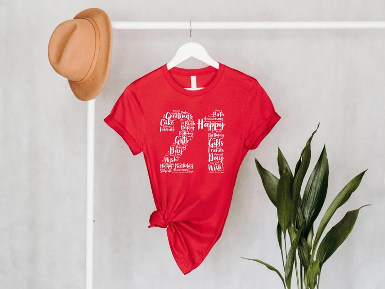 May include: Red short-sleeved t-shirt with a white graphic of the number 27 made up of words related to birthdays, such as "Happy", "Birthday", "Cake", "Gifts", and "Friends".