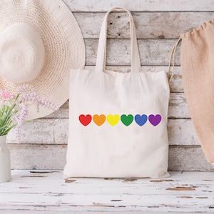 May include: A white canvas tote bag with a row of rainbow-colored hearts printed on the front. The hearts are red, orange, yellow, green, blue, and purple.