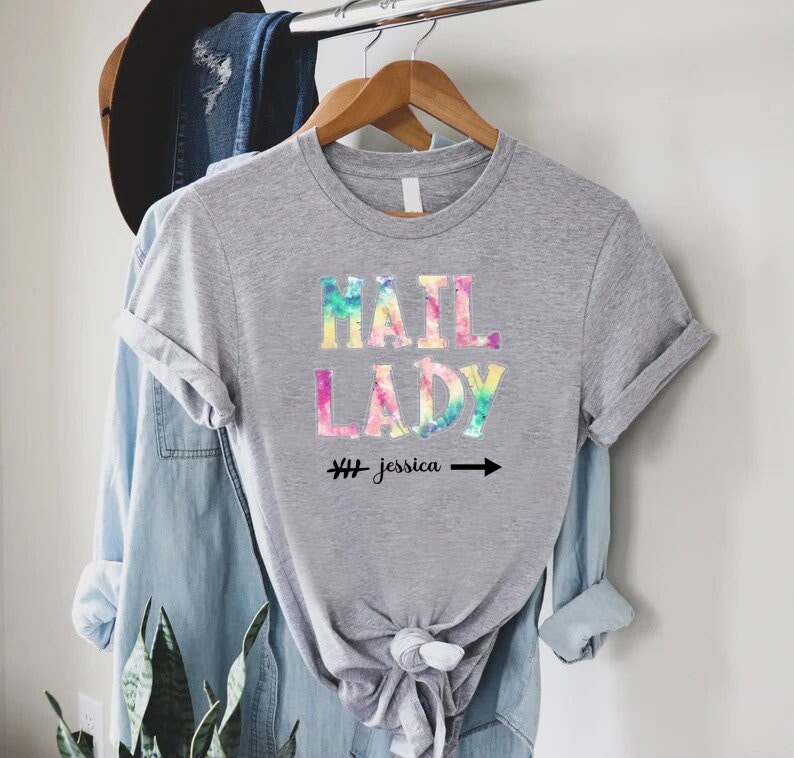 May include: Grey t-shirt with a tie-dye design featuring the text "Mail Lady" and the hashtag "#jessica" with an arrow.