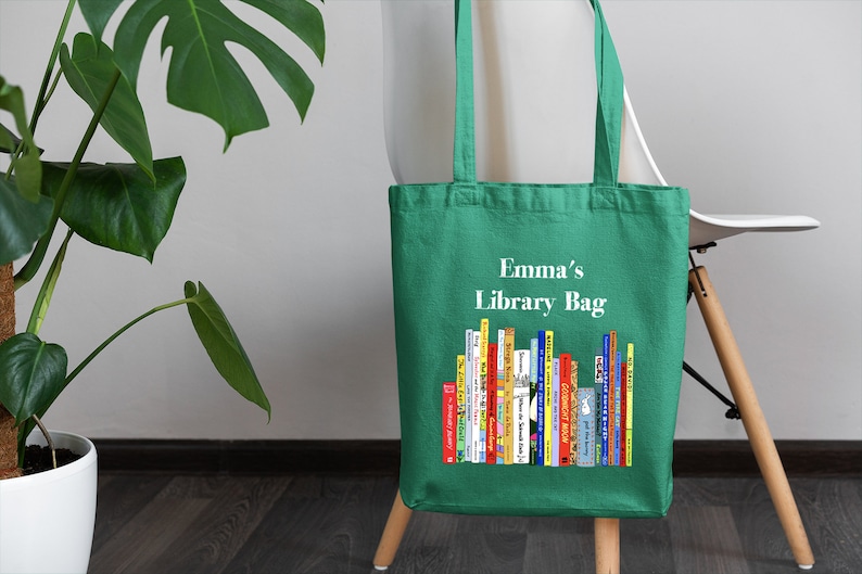 May include: A green tote bag with the text "Emma's Library Bag" and a colorful illustration of books. The bag is made of a sturdy fabric and has long handles for carrying. The bag is sitting on a chair.