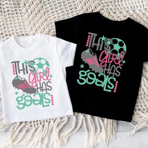 May include: Two white t-shirts with short sleeves and a black t-shirt with short sleeves. The white t-shirts have a graphic design that says "This girl has goals" with a soccer ball and a soccer cleat. The black t-shirt has the same graphic design in pink, green, and gray.