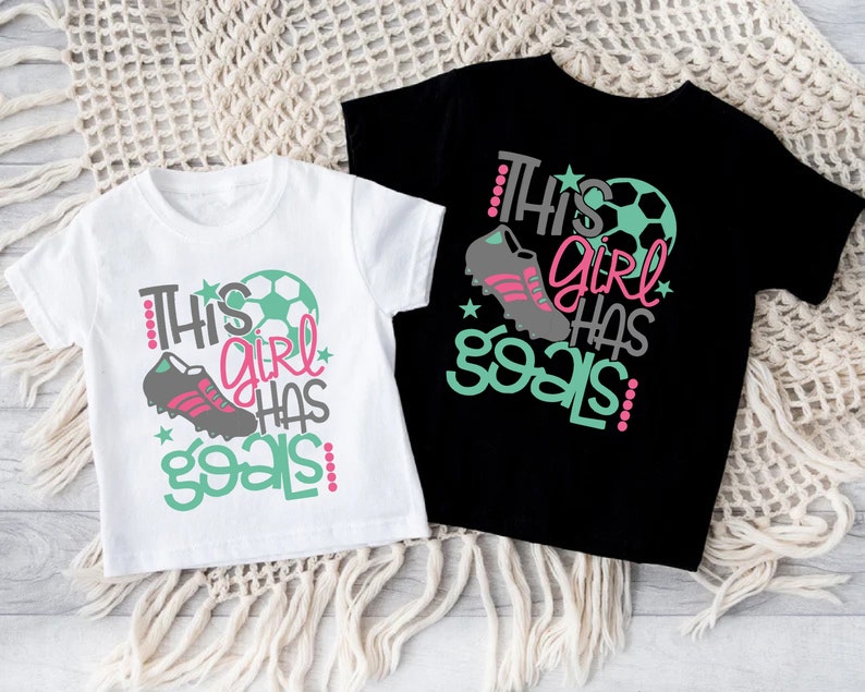 May include: Two white t-shirts with short sleeves and a black t-shirt with short sleeves. The white t-shirts have a graphic design that says "This girl has goals" with a soccer ball and a soccer cleat. The black t-shirt has the same graphic design in pink, green, and gray.