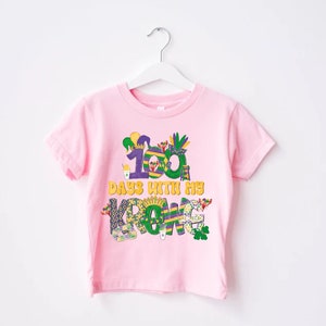 May include: Pink t-shirt featuring a colourful graphic that reads "100 days with my crew". The graphic incorporates Mardi Gras themed elements such as beads, masks, and a king cake.