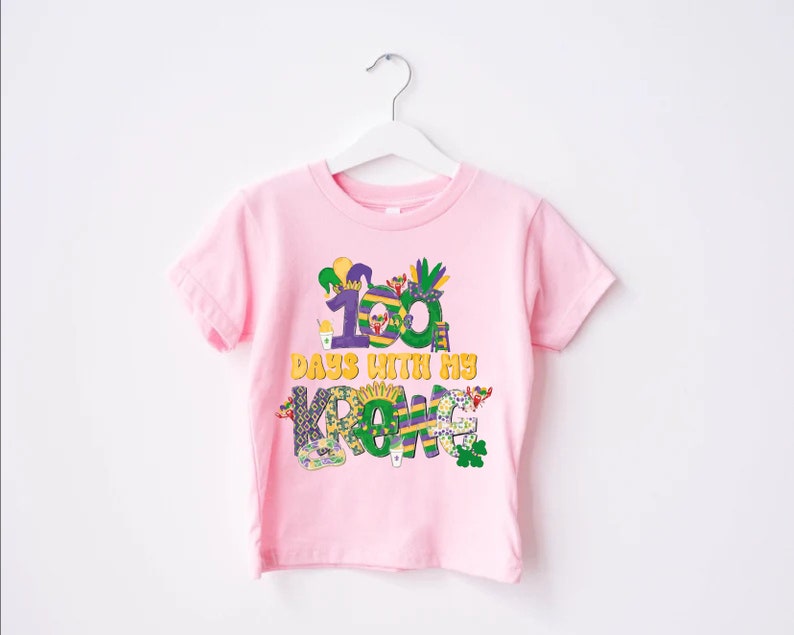 May include: Pink t-shirt featuring a colourful graphic that reads "100 days with my crew". The graphic incorporates Mardi Gras themed elements such as beads, masks, and a king cake.
