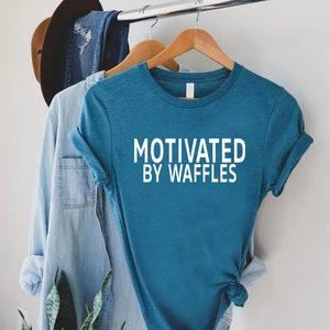 K&ouml;nnte beinhalten: Ein t&uuml;rkisfarbenes T-Shirt mit wei&szlig;em Text, der "Motivated by Waffles" lautet. Das Shirt h&auml;ngt an einem Holzb&uuml;gel.