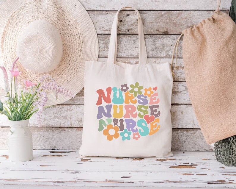 Puede incluir: Una bolsa de lona blanca con un dise&ntilde;o gr&aacute;fico colorido que dice "NURSE" en una fuente retro con flores y corazones.