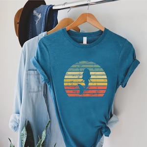 May include: A teal blue t-shirt with a vintage-style sunset graphic featuring a mermaid silhouette. The graphic has a distressed look with yellow, orange, and red stripes.
