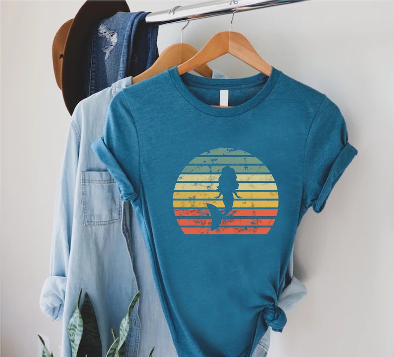 May include: A teal blue t-shirt with a vintage-style sunset graphic featuring a mermaid silhouette. The graphic has a distressed look with yellow, orange, and red stripes.