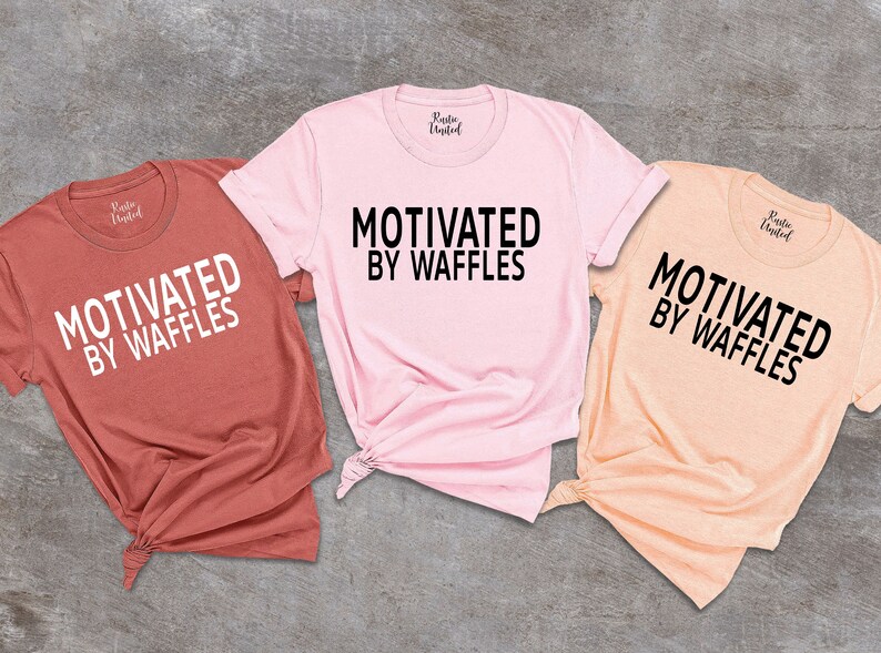 K&ouml;nnte beinhalten: Drei T-Shirts mit dem Text "Motivated by Waffles" in schwarzer Schrift auf wei&szlig;em Hintergrund. Die Shirts sind in verschiedenen Farben: Rost, Pink und Pfirsich.