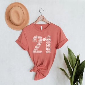 May include: A rust-colored t-shirt with a white graphic design of the number 21 made up of words related to birthdays, such as "Happy", "Birthday", "Cake", "Friends", and "Gifts".