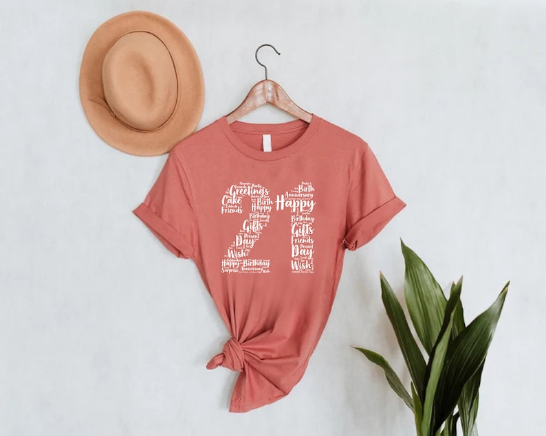 May include: A rust-colored t-shirt with a white graphic design of the number 21 made up of words related to birthdays, such as "Happy", "Birthday", "Cake", "Friends", and "Gifts".