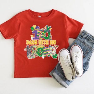 May include: Red short-sleeved t-shirt featuring a colourful graphic that reads "100 Days with my Crew". The graphic is in the style of Mardi Gras decorations. The shirt is paired with blue jeans and white trainers.