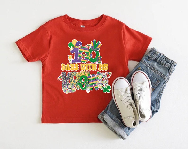 May include: Red short-sleeved t-shirt featuring a colourful graphic that reads "100 Days with my Crew". The graphic is in the style of Mardi Gras decorations. The shirt is paired with blue jeans and white trainers.