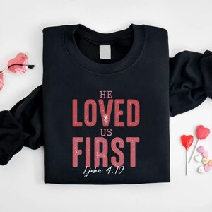 Puede incluir: Sudadera negra con un gr&aacute;fico rojo que dice "He Loved Us First 1 John 4:19".