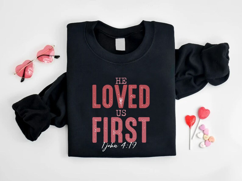 Puede incluir: Sudadera negra con un gr&aacute;fico rojo que dice "He Loved Us First 1 John 4:19".