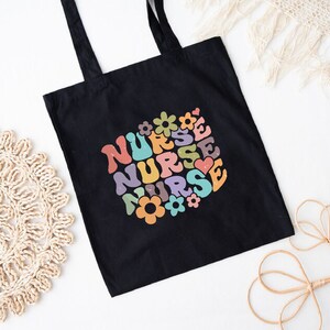 Puede incluir: Bolsa de tela negra con un dise&ntilde;o retro colorido que dice "NURSE" varias veces con flores y corazones alrededor del texto.
