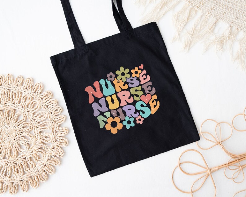 Puede incluir: Bolsa de tela negra con un dise&ntilde;o retro colorido que dice "NURSE" varias veces con flores y corazones alrededor del texto.