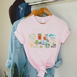 Puede incluir: Camiseta rosa con un dise&ntilde;o gr&aacute;fico de varios iconos ambientales, incluyendo una bombilla, &aacute;rboles, un globo terr&aacute;queo y el texto "Think Green", "Save Our Seas", "This is my home too", "Do Not Use Plastic", "Save Energy", y "More Trees".
