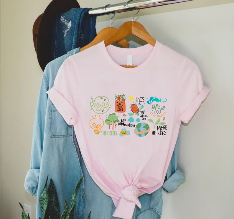 Puede incluir: Camiseta rosa con un dise&ntilde;o gr&aacute;fico de varios iconos ambientales, incluyendo una bombilla, &aacute;rboles, un globo terr&aacute;queo y el texto "Think Green", "Save Our Seas", "This is my home too", "Do Not Use Plastic", "Save Energy", y "More Trees".