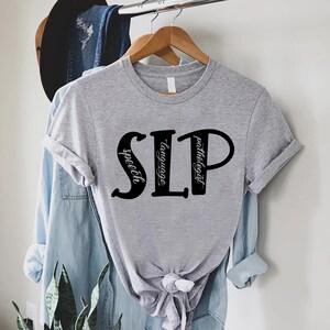 Puede incluir: Camiseta gris con texto negro que dice "SLP" con las palabras "speech", "language" y "pathologist" escritas verticalmente dentro de las letras.