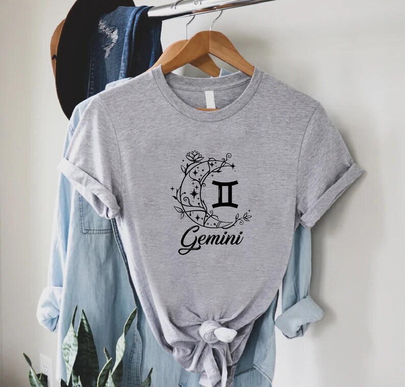 Puede incluir: Una camiseta gris con un gr&aacute;fico negro del signo zodiacal G&eacute;minis dentro de una luna creciente con detalles florales. El texto "Gemini" est&aacute; escrito debajo de la luna.