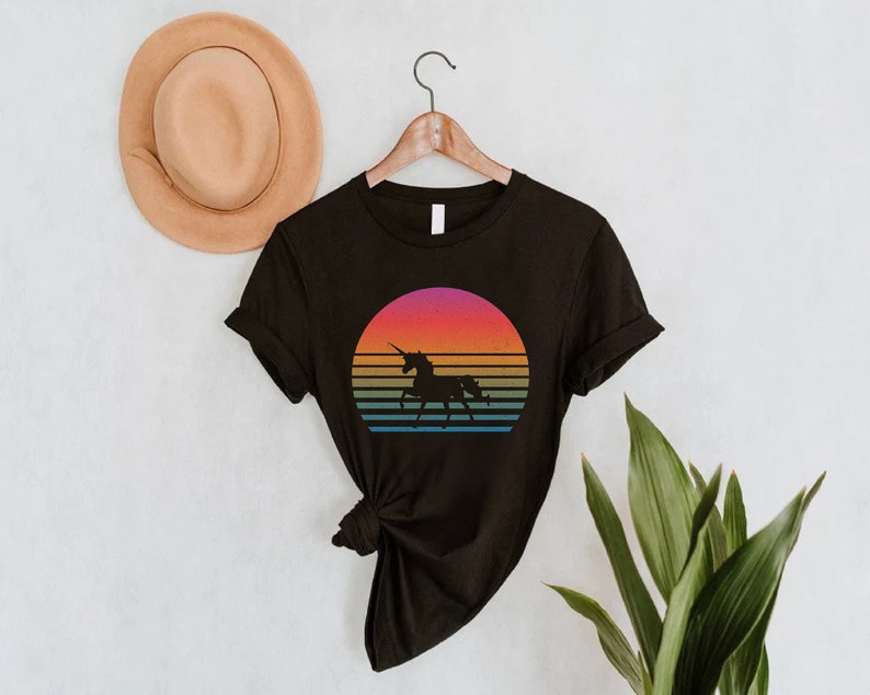 K&ouml;nnte beinhalten: Schwarzes T-Shirt mit einem Retro-Sonnenuntergangsdesign und einer Einhorn-Silhouette. Der Sonnenuntergang hat B&auml;nder in Orange, Rosa, Gelb und Blau. Das T-Shirt h&auml;ngt an einem Holzb&uuml;gel.