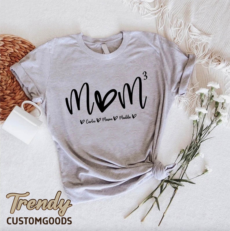 May include: Gray t-shirt with the text "MOM" in black with a heart replacing the "O". The text "3" is to the right of the "M". Below the text is "Carlee, Mason, Maddie" with a heart before and after each name.