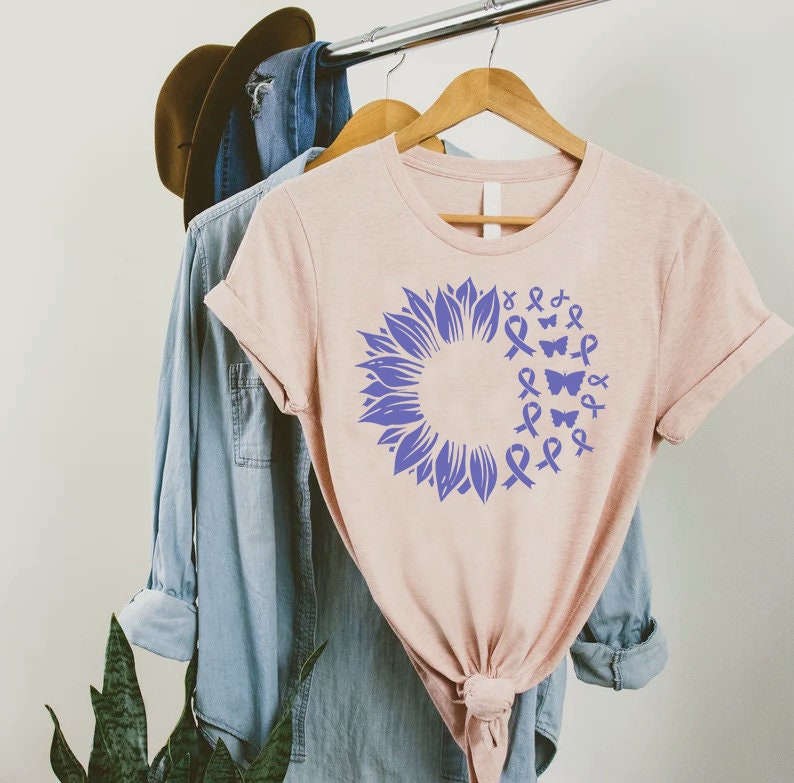 May include: A light pink t-shirt featuring a purple sunflower design. The sunflower is made up of purple ribbons and butterflies.