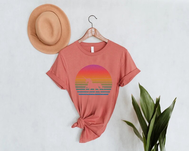 K&ouml;nnte beinhalten: Ein korallenfarbenes T-Shirt mit einem Retro-Design, das eine Einhorn-Silhouette vor einem farbenfrohen Sonnenuntergang zeigt. Das Design umfasst horizontale Streifen in Orange-, Gelb-, Gr&uuml;n- und Blaut&ouml;nen. Ein hellbrauner Hut h&auml;ngt an der Wand &uuml;ber dem Shirt.