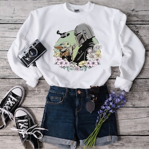 May include: White sweatshirt featuring a graphic print of a Mandalorian character and a Grogu character with flowers. The sweatshirt is styled with blue denim shorts, black trainers, and a bunch of purple flowers.
