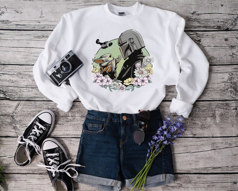 May include: White sweatshirt featuring a graphic print of a Mandalorian character and a Grogu character with flowers. The sweatshirt is styled with blue denim shorts, black trainers, and a bunch of purple flowers.