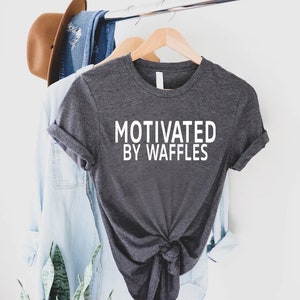 K&ouml;nnte beinhalten: Ein dunkelgraues T-Shirt mit dem Aufdruck "Motivated by Waffles" in Wei&szlig; auf der Vorderseite.