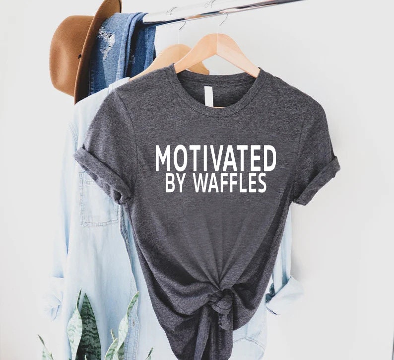 K&ouml;nnte beinhalten: Ein dunkelgraues T-Shirt mit dem Aufdruck "Motivated by Waffles" in Wei&szlig; auf der Vorderseite.