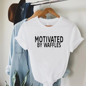 K&ouml;nnte beinhalten: Wei&szlig;es T-Shirt mit schwarzem Text, der "Motiviert durch Waffeln" lautet.