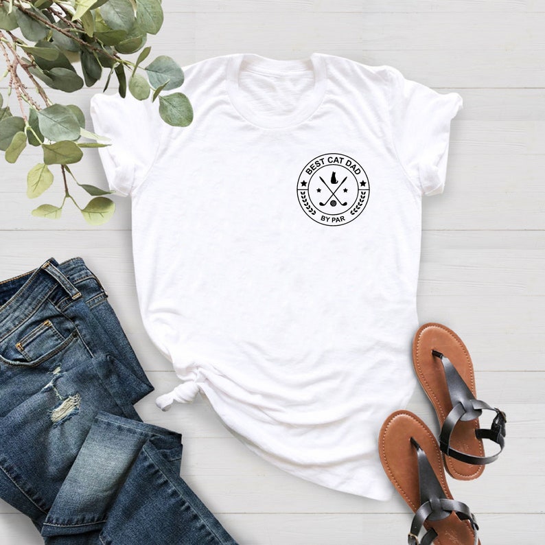 May include: White t-shirt with a circular graphic that reads "BEST CAT DAD BY PAR" in black. The graphic includes a cat silhouette, crossed golf clubs, and stars. The shirt is paired with blue jeans and brown sandals.