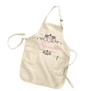 Puede incluir: Delantal color crema con dos bolsillos delanteros y un portal&aacute;pices. El delantal presenta el nombre "Sarah" en escritura rosa, con adornos decorativos negros y un dise&ntilde;o estilizado en la parte superior. Correa ajustable para el cuello.