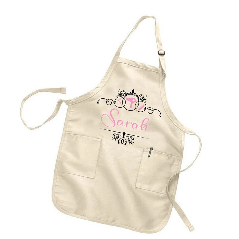 Puede incluir: Delantal color crema con dos bolsillos delanteros y un portal&aacute;pices. El delantal presenta el nombre "Sarah" en escritura rosa, con adornos decorativos negros y un dise&ntilde;o estilizado en la parte superior. Correa ajustable para el cuello.