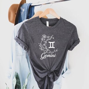Puede incluir: Una camiseta gris oscuro con un gr&aacute;fico blanco de una luna creciente con enredaderas y estrellas, el s&iacute;mbolo del zod&iacute;aco G&eacute;minis y la palabra "G&eacute;minis".