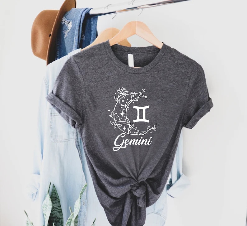 Puede incluir: Una camiseta gris oscuro con un gr&aacute;fico blanco de una luna creciente con enredaderas y estrellas, el s&iacute;mbolo del zod&iacute;aco G&eacute;minis y la palabra "G&eacute;minis".