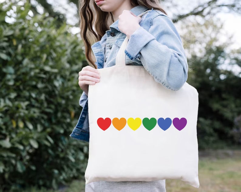 May include: A white tote bag with a rainbow of hearts printed on the front. The hearts are red, orange, yellow, green, blue, and purple.