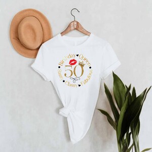 May include: White t-shirt with a gold glitter design that says "Birthday Queen 50 Sassy Classy Fabulous" with a red lip and a gold crown.