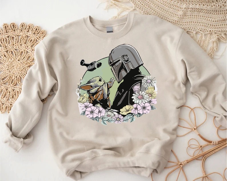 May include: A beige sweatshirt featuring a printed design of a Mandalorian character and a Grogu character surrounded by flowers.