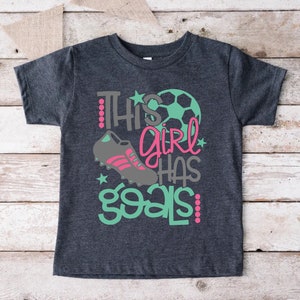 May include: A dark gray t-shirt with the text "This girl has goals" in pink, gray, and teal. A soccer ball and a soccer cleat are also featured on the shirt.