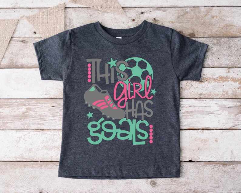 May include: A dark gray t-shirt with the text "This girl has goals" in pink, gray, and teal. A soccer ball and a soccer cleat are also featured on the shirt.