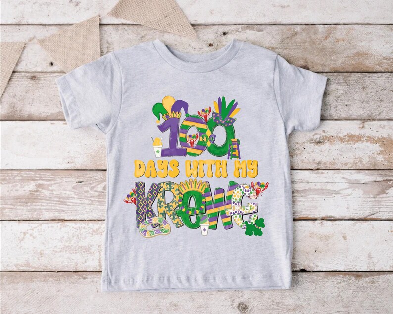 May include: A grey t-shirt with a colourful design featuring the text "100 Days With My Krewe". The design includes Mardi Gras themed elements like beads, a jester hat, and a king cake.