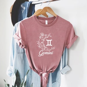 Puede incluir: Una camiseta rosa con un gr&aacute;fico blanco de una luna creciente con enredaderas florales y el s&iacute;mbolo del zod&iacute;aco G&eacute;minis. El texto "G&eacute;minis" est&aacute; escrito debajo del s&iacute;mbolo.