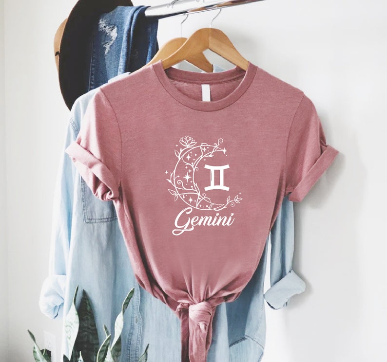 Puede incluir: Una camiseta rosa con un gr&aacute;fico blanco de una luna creciente con enredaderas florales y el s&iacute;mbolo del zod&iacute;aco G&eacute;minis. El texto "G&eacute;minis" est&aacute; escrito debajo del s&iacute;mbolo.