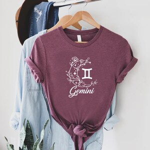 Puede incluir: Una camiseta color granate con un gr&aacute;fico blanco de una luna creciente con enredaderas florales, estrellas y el s&iacute;mbolo del zod&iacute;aco G&eacute;minis. El texto "G&eacute;minis" est&aacute; escrito debajo del s&iacute;mbolo.